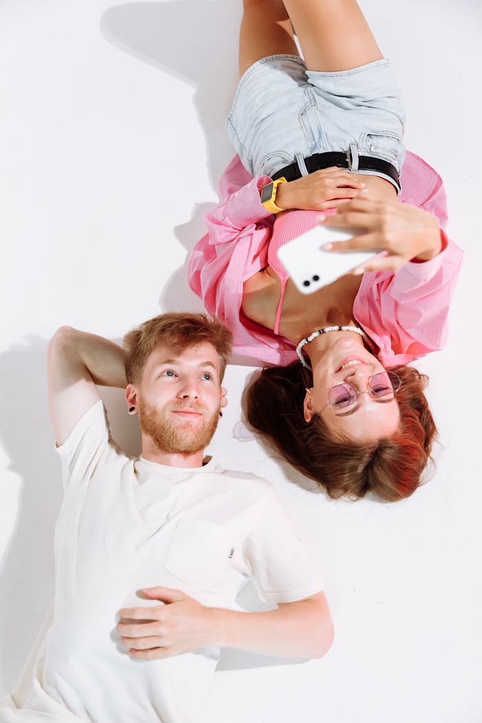 A cheerful young couple lying on a white floor, capturing a fun selfie with a smartphone.